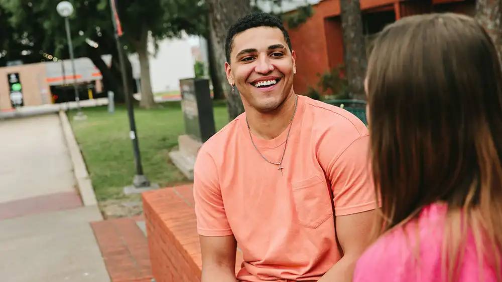 ​​A student in a peach-colored shirt sits on a red brick bench outdoors, smiling and talking to another student in a pink top whose back is to the camera. Trees, a lawn, and apartment buildings are visible in the background, suggesting a college friendly setting.​ 