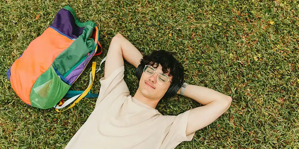 ​​Student lying on grass with eyes closed, wearing headphones and a beige shirt, relaxing next to a colorful backpack.​ 