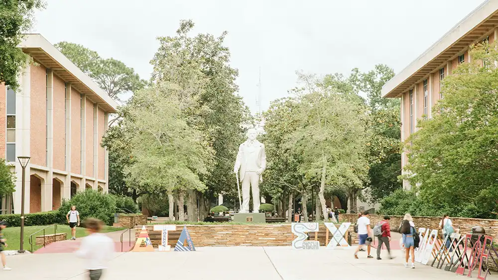 ​​A bright, outdoor scene from the sidelines of the Frank Parker Plaza. A statue of Sam Houston himself stands in the middle of the courtyard, surrounded by lush green trees. Various fraternity and sorority letters and signs are displayed. Students walk and gather in small groups, some heading towards buildings, while others pass by.​ 
