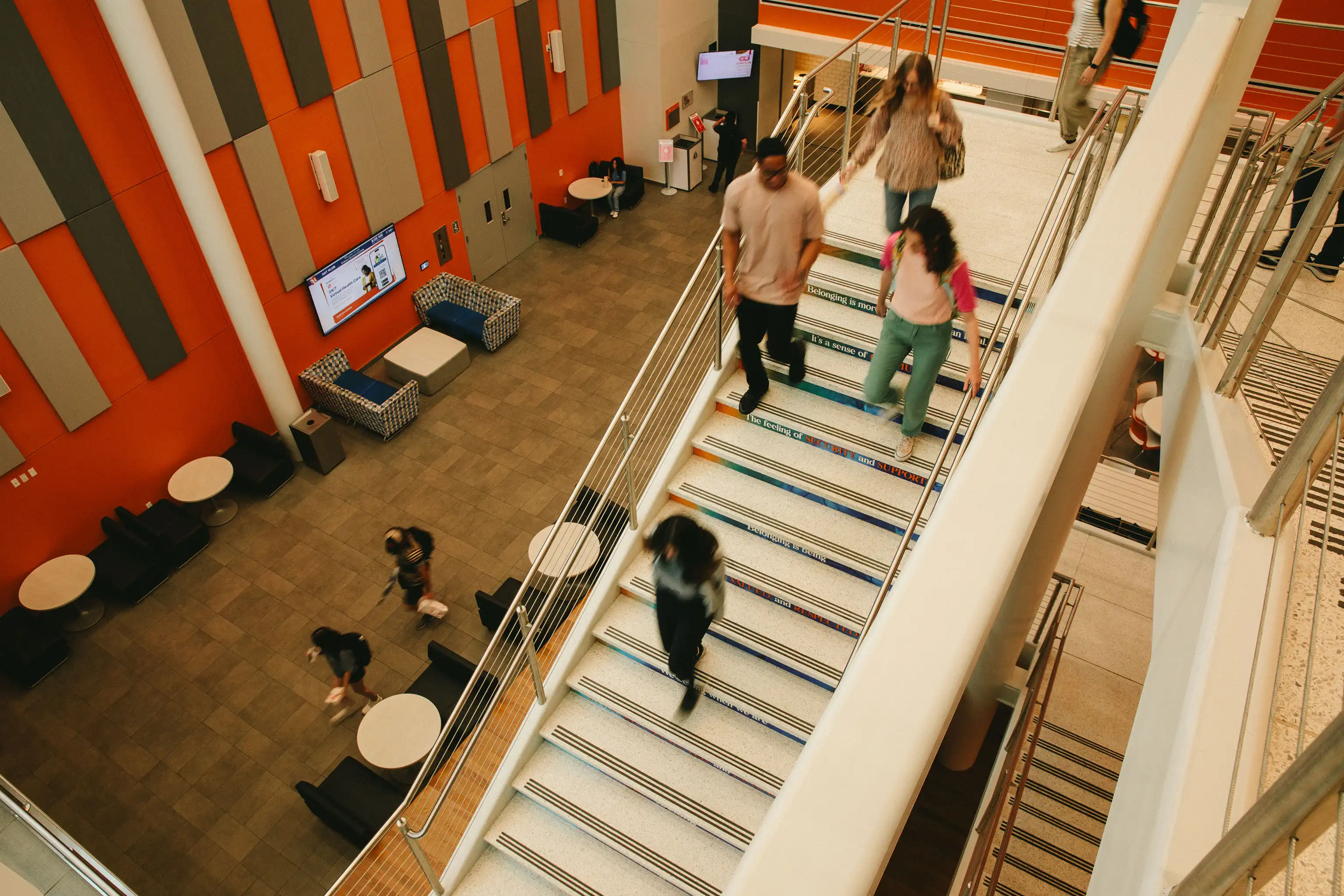 ​​Students walk up and down a brightly lit staircase inside the Lowman Student Center with orange and gray wall panels, lounge seating, and round tables below.​ 
