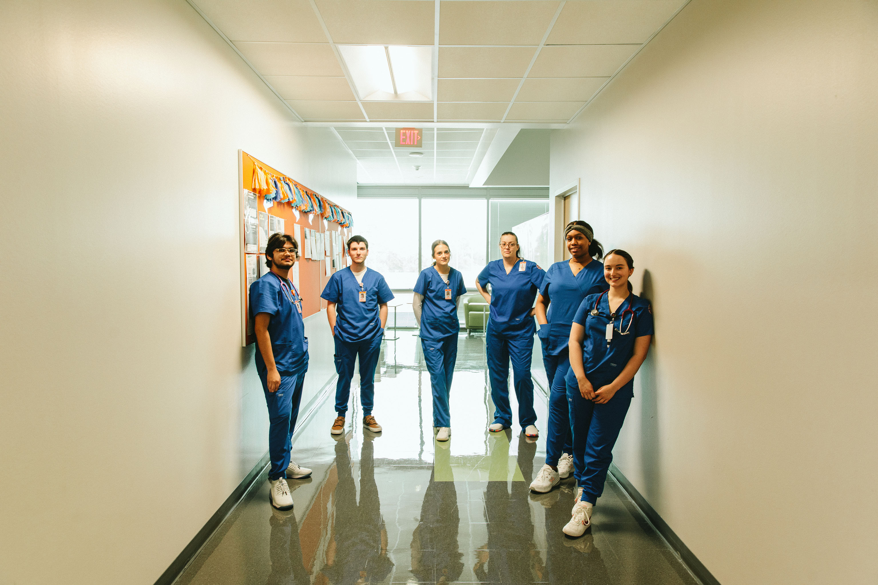 ​​Six individuals in blue medical scrubs and white shoes stand in a semi-circle in a bright hallway, facing the camera. The hallway features light-colored walls, a reflective floor, and a decorated bulletin board on the left. An exit sign and large windows in the background allow natural light into the space.​ 